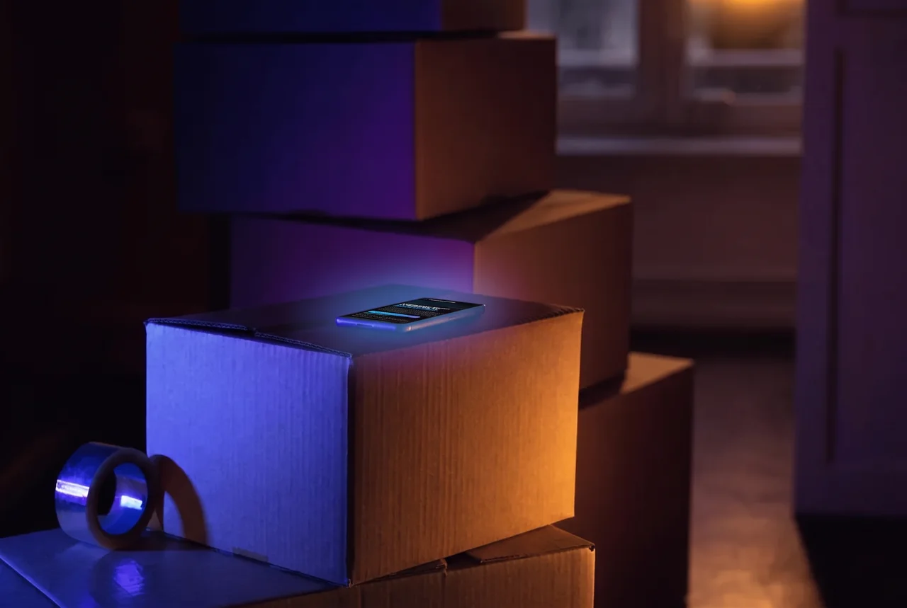 Cardboard moving boxes stacked in an empty living room at dusk, a phone glowing on top of the nearest box beside a roll of packing tape.