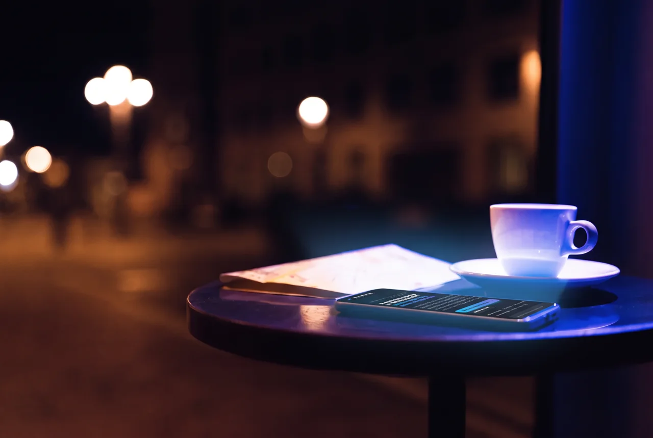 A phone on a small café table at night with a folded street map and an espresso cup, city lights blurred in the window.