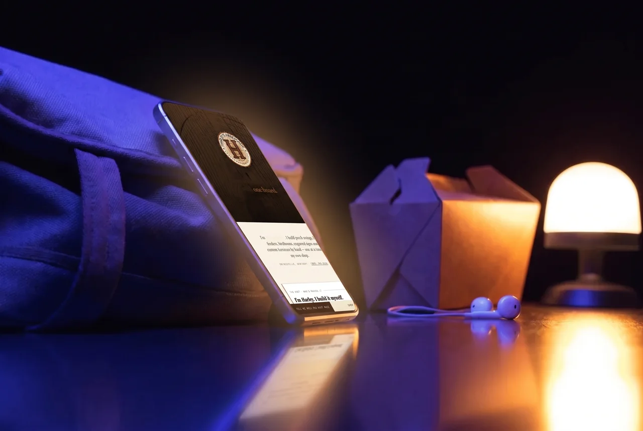 A phone leaning against a canvas backpack on a dorm desk with a takeout box, earbuds and a small lamp, late at night.